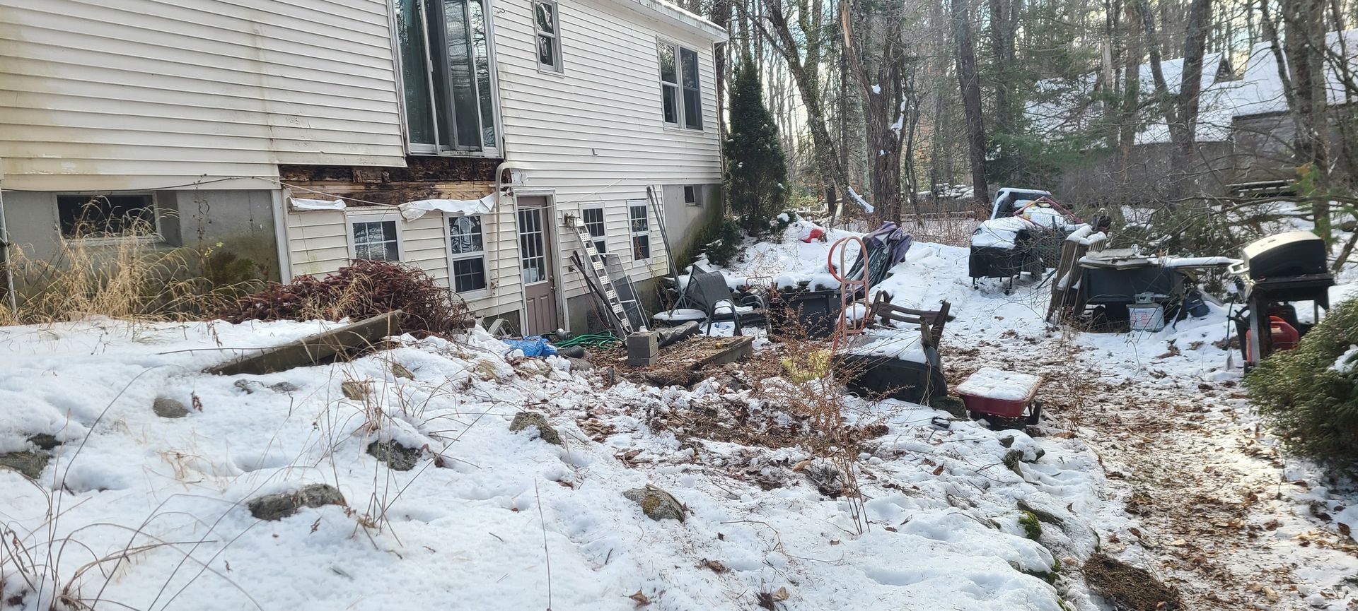 Snow-covered yard beside a house with scattered debris and a few chairs near leafless trees