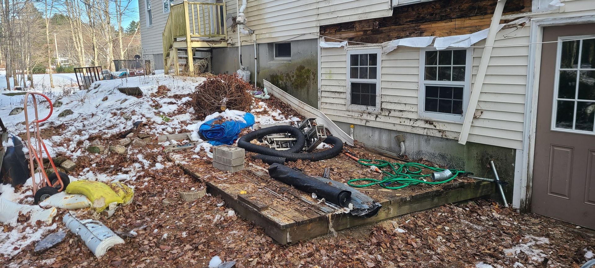 Flood-damaged exterior with debris, torn siding, broken windows, and an overturned motorcycle beside a house