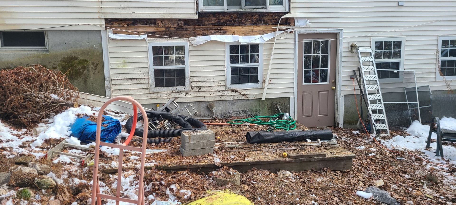 Snowy backyard with scattered debris and tools beside a house’s damaged foundation