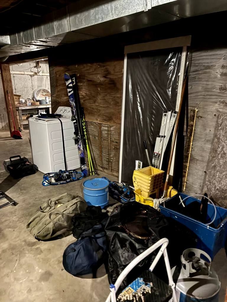 Cluttered basement storage area with bags, a blue bin, folded chairs, and a plastic-covered doorway