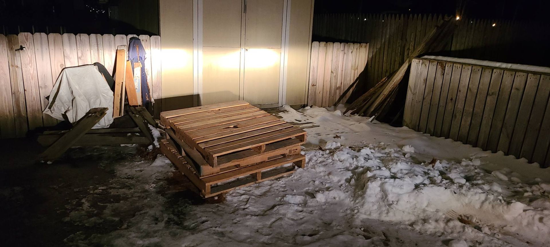 Snowy backyard with stacked wooden pallets, leaning boards, and a lit fence at night