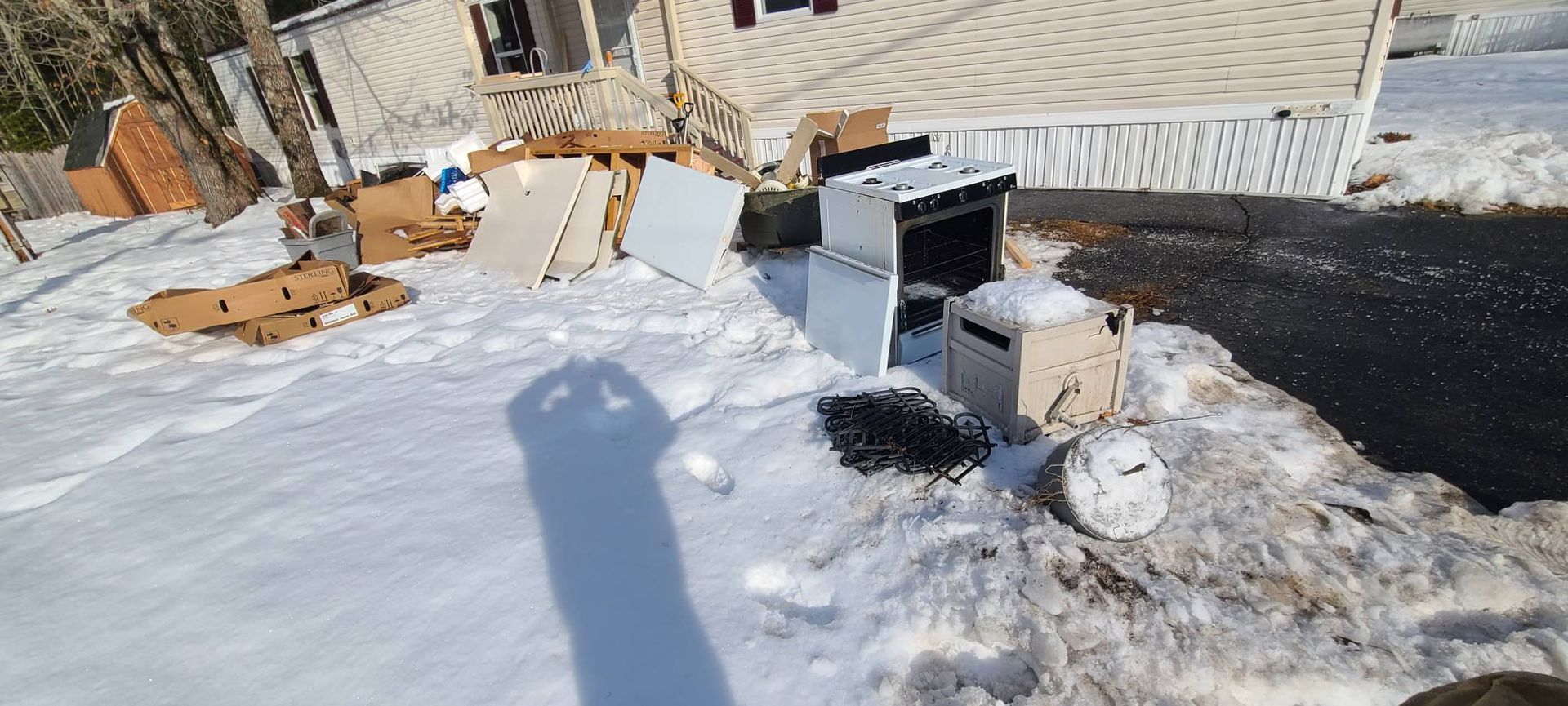 Snowy backyard with scattered cardboard, a grill, and a round metal dish beside a fence