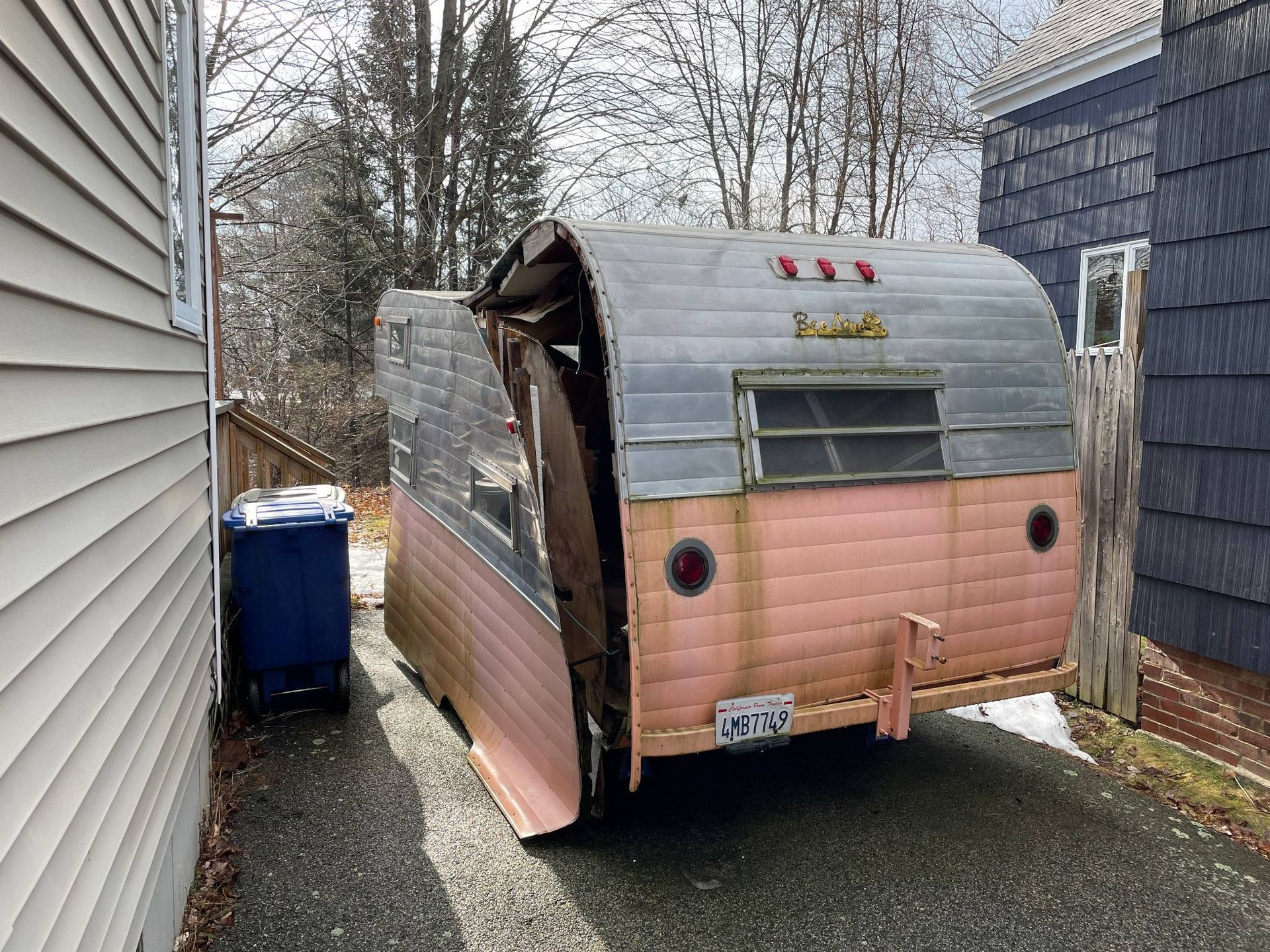 Damaged pink and silver camper trailer parked between two houses in a narrow side yard