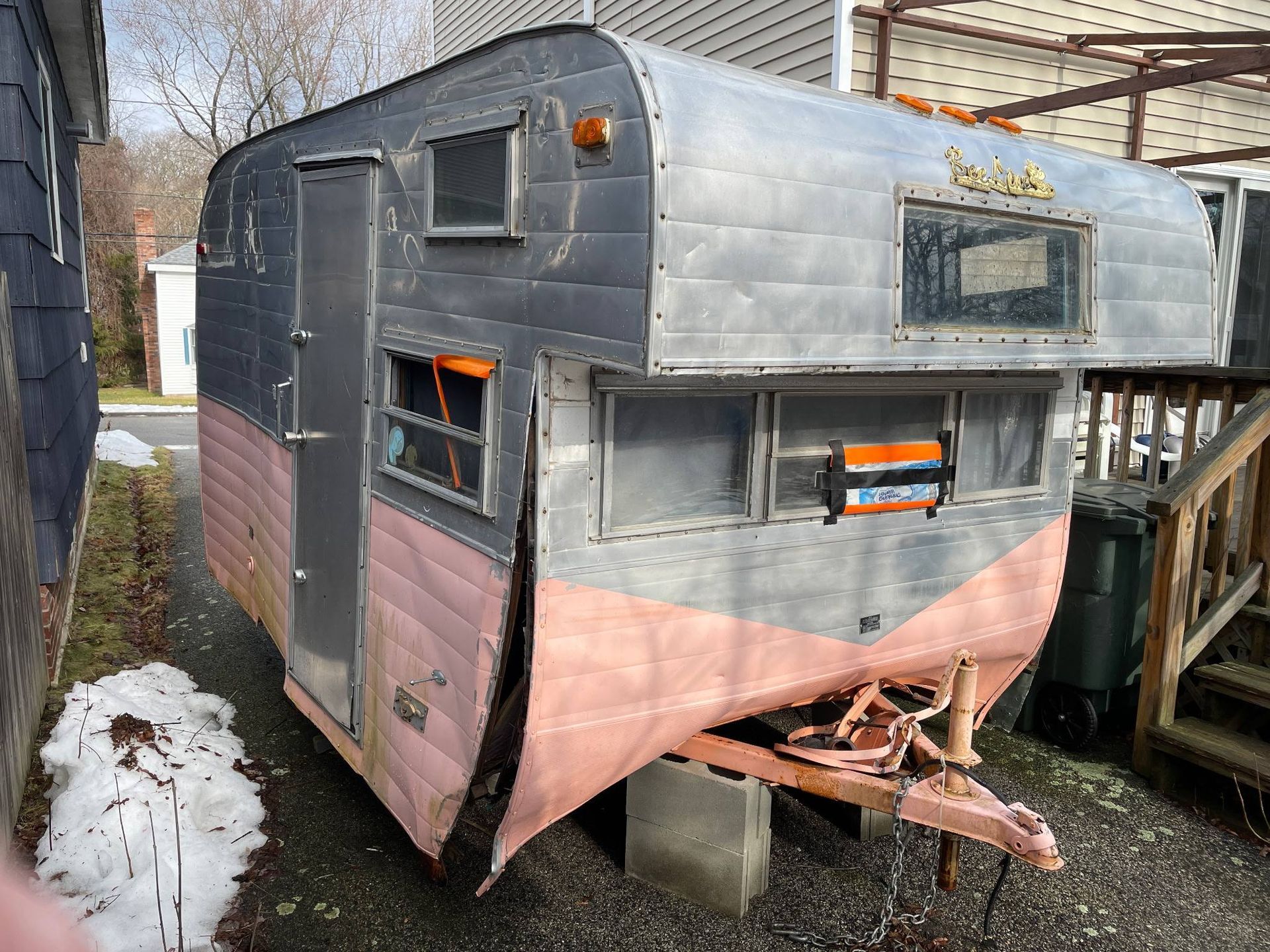 Old silver and pink camper trailer with orange windows, parked beside a house on a driveway.