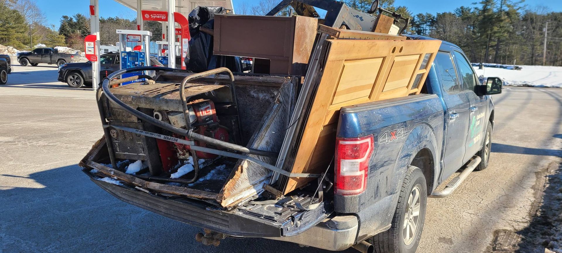 Blue pickup truck with a damaged bed piled with debris, parked on a snowy roadside outdoors