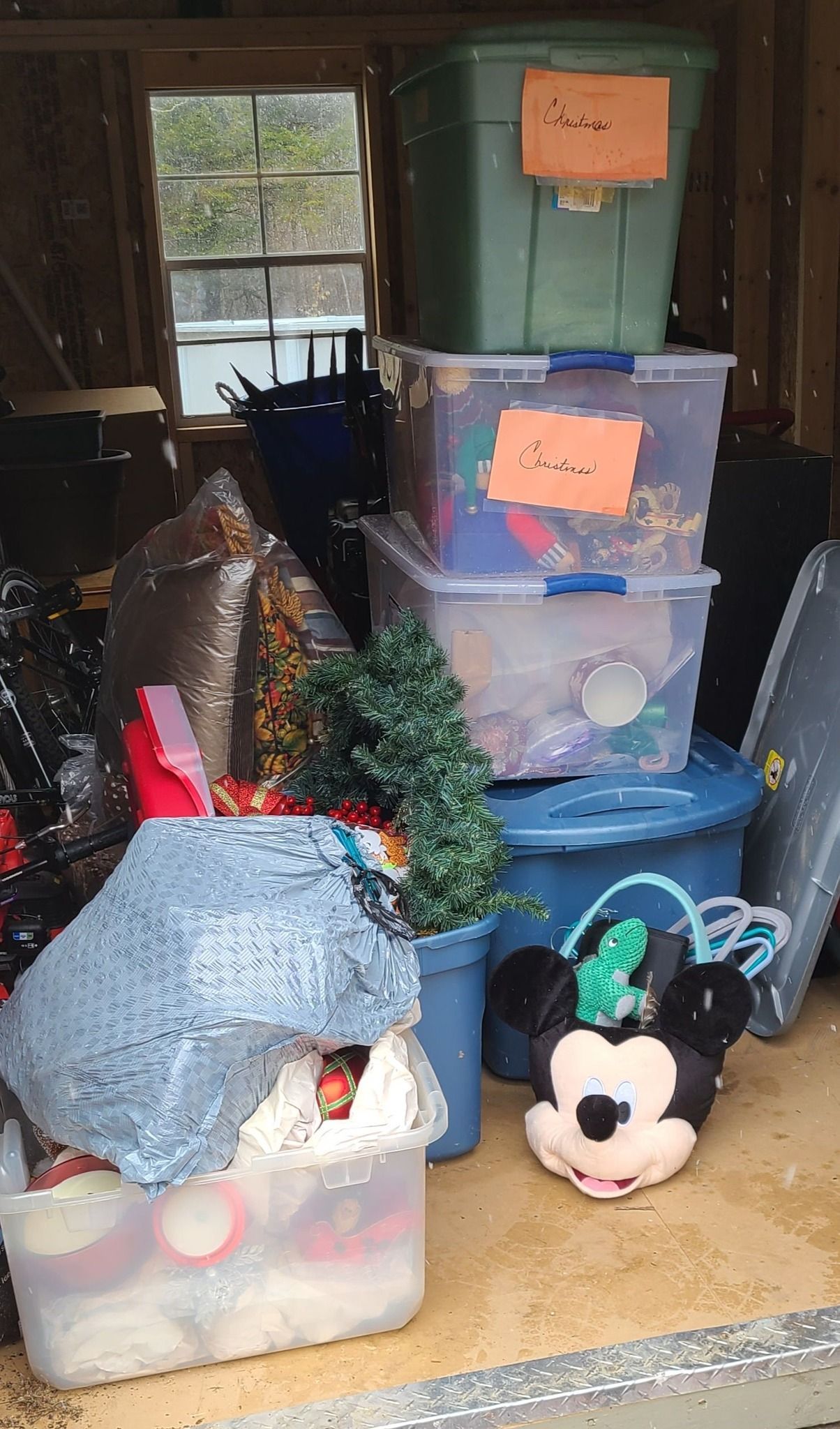 Stacked clear plastic storage bins in a dim room, with holiday decorations and a Mickey Mouse plush on the floor.