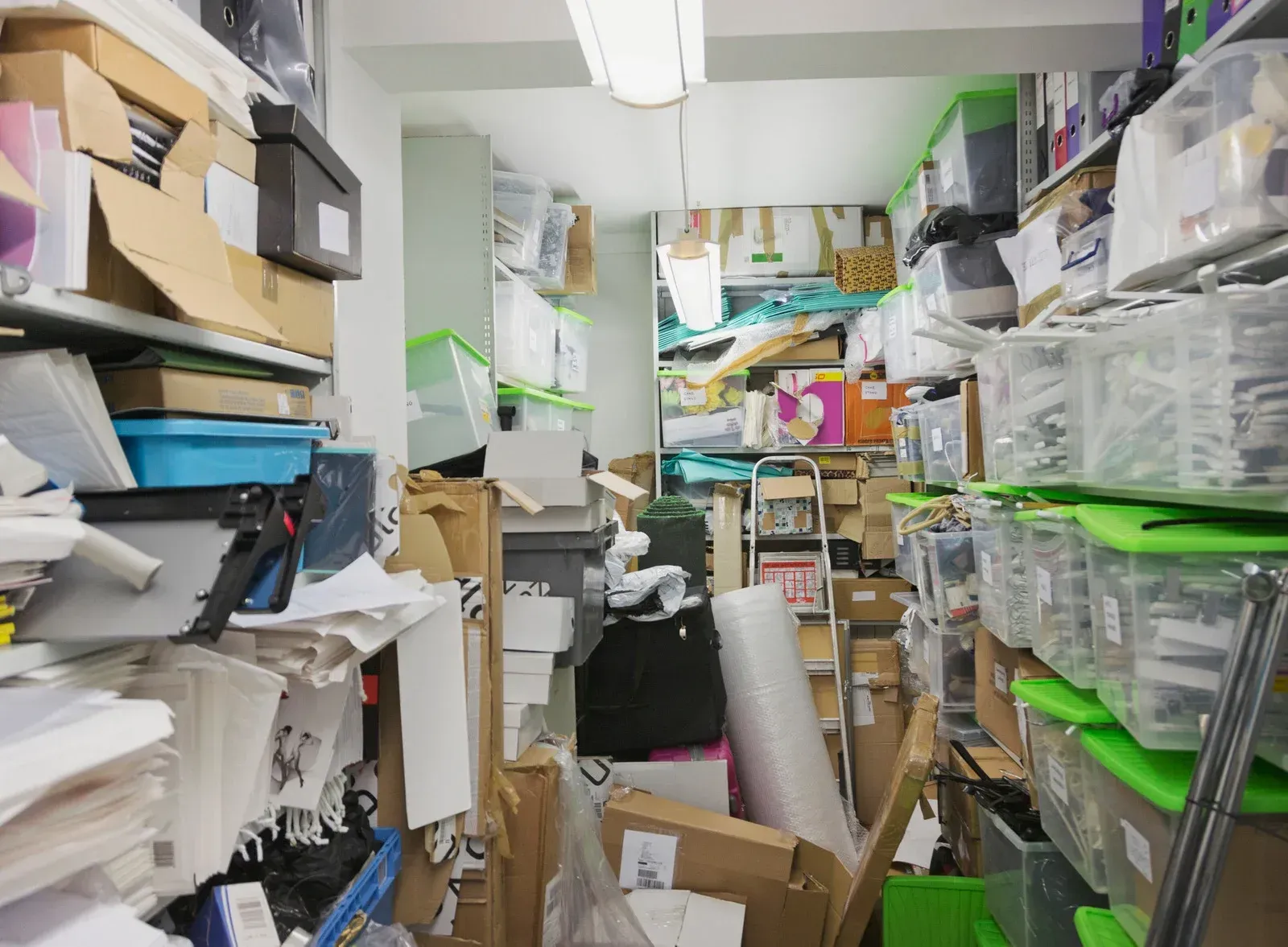 Cluttered storage room packed with boxes, plastic bins, papers, and assorted supplies.