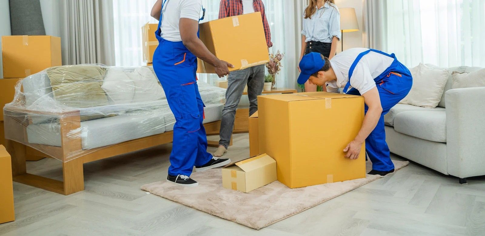 Movers in blue uniforms carrying boxes in a bright living room with wrapped furniture