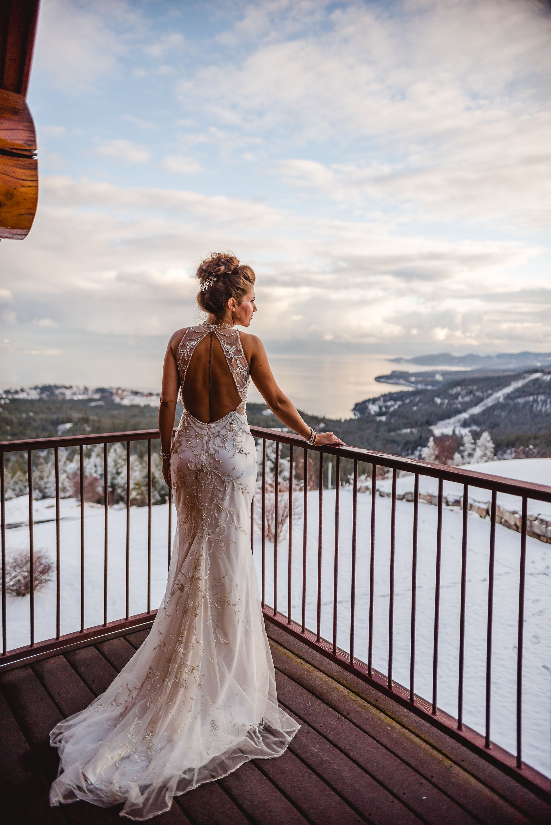 Woman in a white lace gown standing on a snowy balcony overlooking mountains at sunset