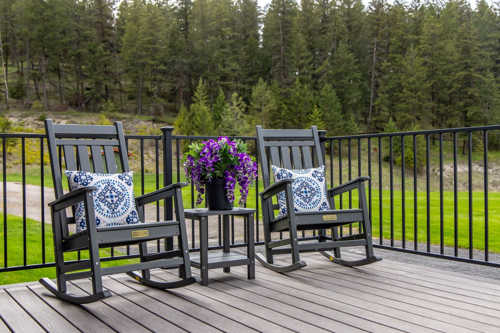 Two gray rocking chairs with floral pillows on a deck beside a table of purple flowers, overlooking trees and water