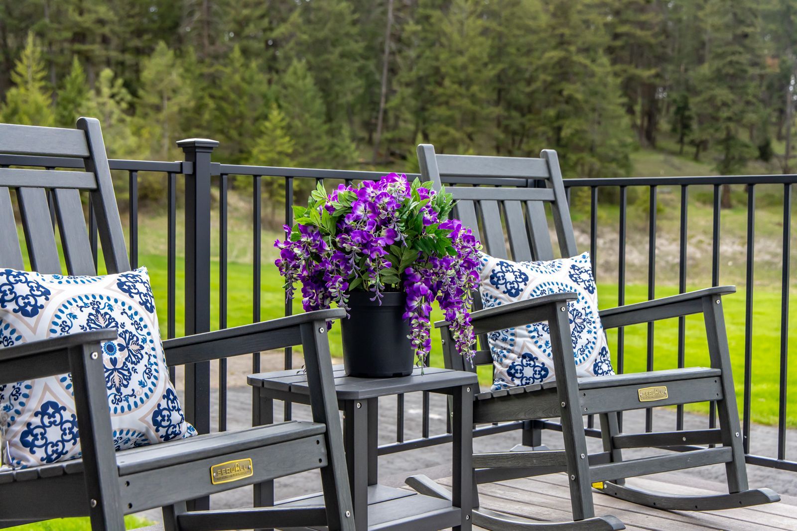 Patio chairs with blue patterned pillows beside a black table of purple flowers on a deck by a grassy yard and trees