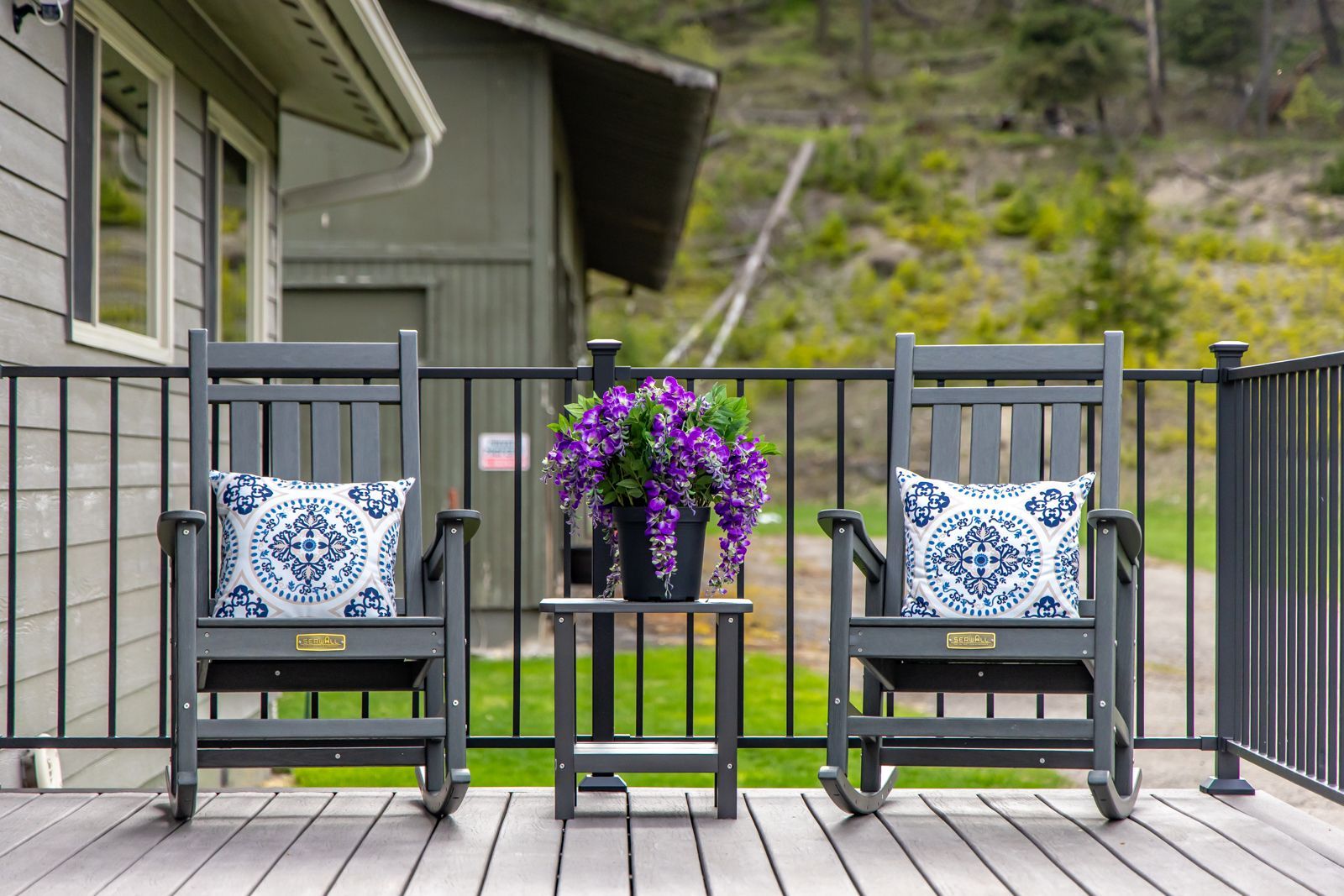Two black chairs with blue patterned pillows flank a small table with purple flowers on a deck.