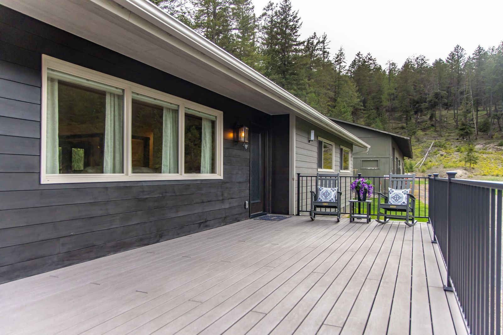 Modern cabin deck with dark siding, large windows, and outdoor seating in a wooded area