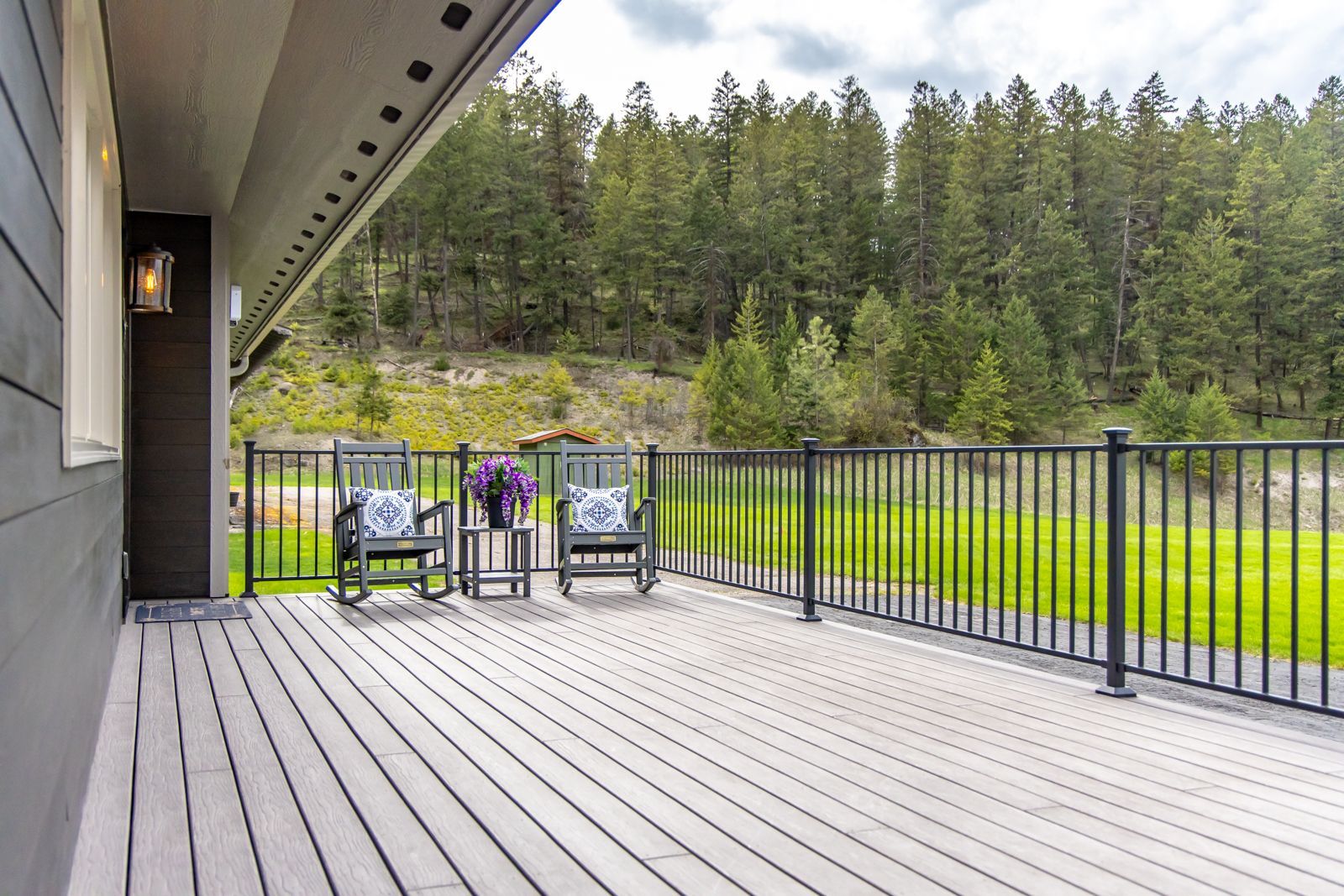 Covered deck with white flooring, black railings, and chairs overlooking a grassy field and forest.