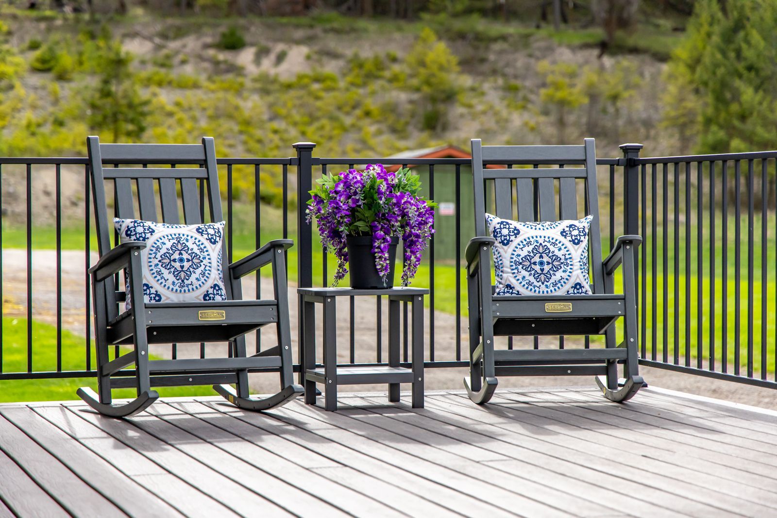 Two black rocking chairs with blue patterned cushions and a purple flower pot on a wooden deck by a black fence