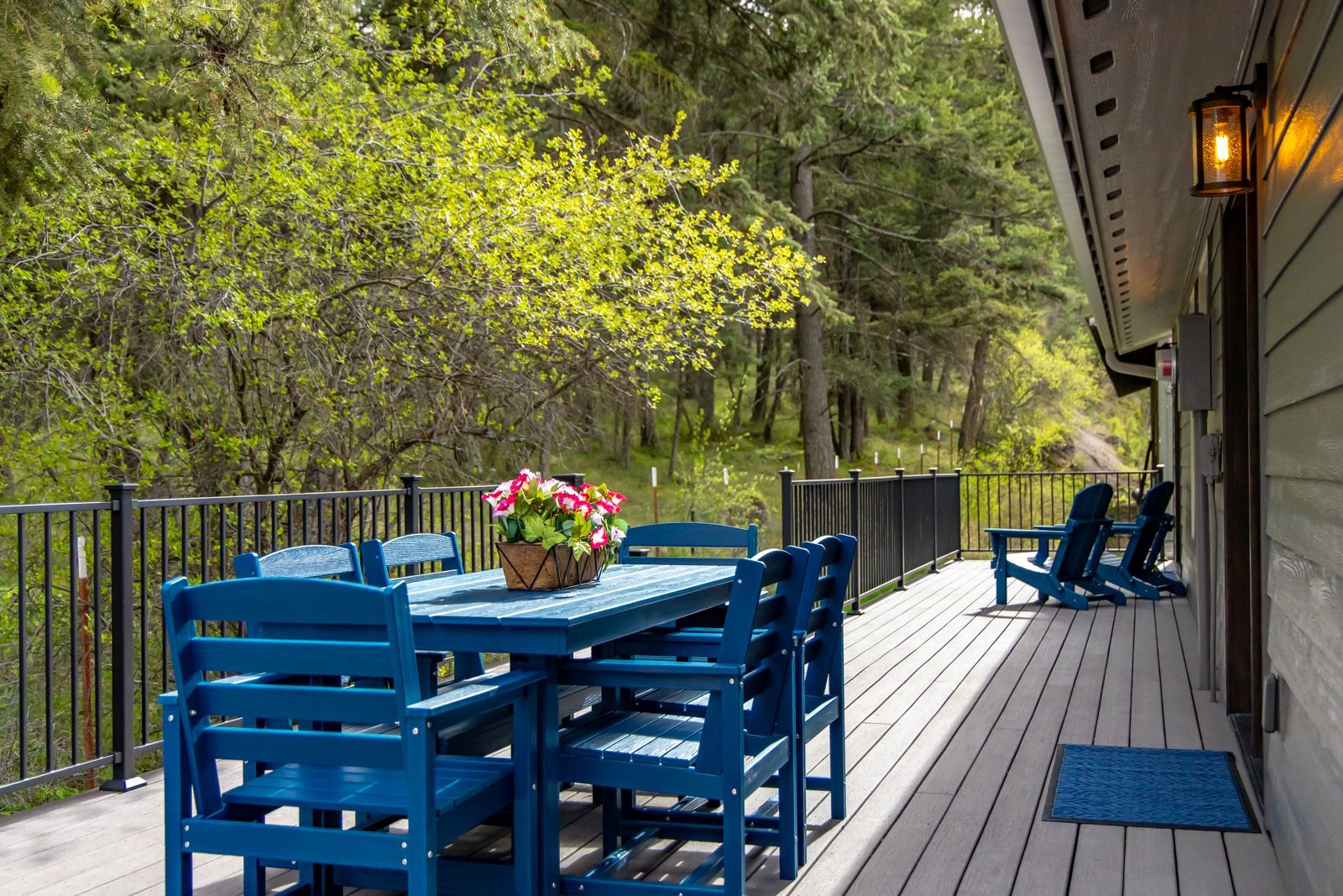 Blue patio chairs and table on a wooden deck beside a forested walkway, with lanterns and a river view