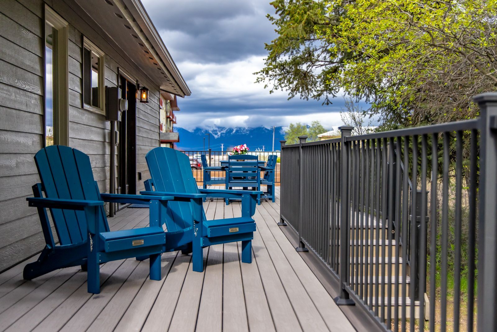 Blue Adirondack chairs on a lakeside deck beside a house, with railing, trees, and cloudy sky.