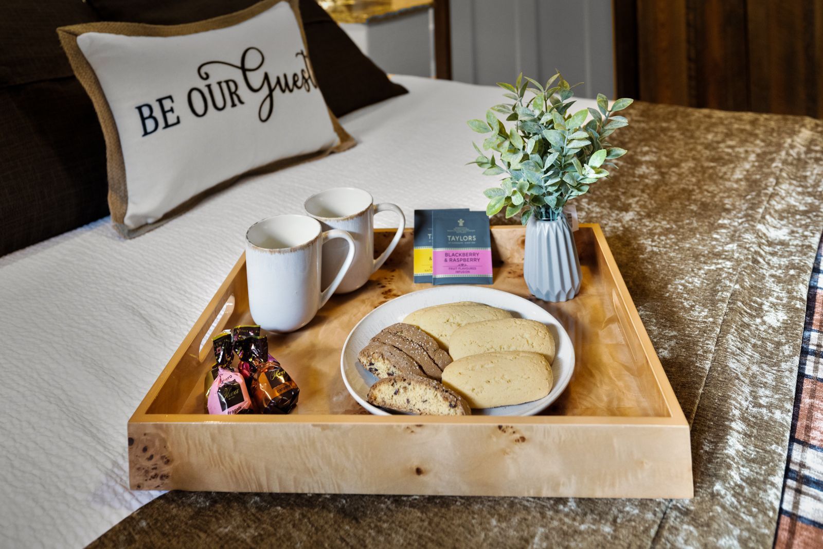 Breakfast tray with tea, cookies, flowers, and a book on a bed with a “Be Our Guest” pillow