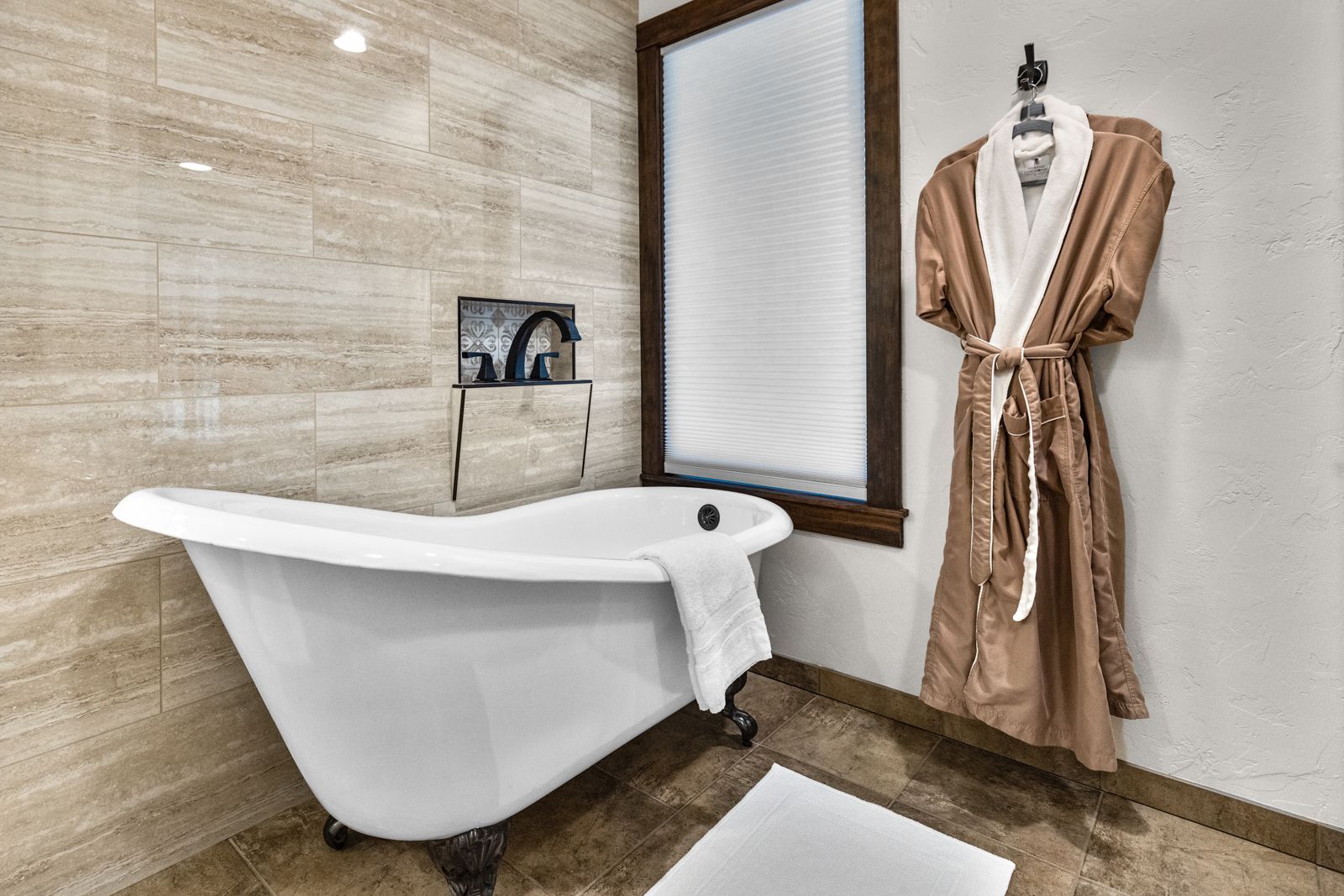 Modern bathroom with a freestanding white tub, beige tile walls, and a robe hanging by the window.