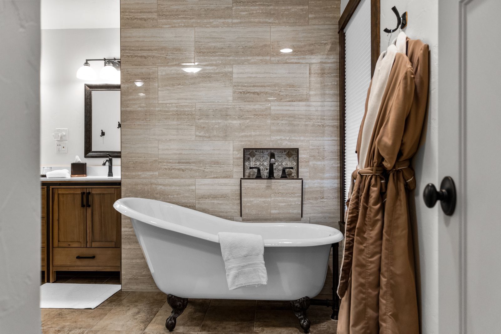 Modern bathroom with freestanding tub, beige tile wall, vanity mirror, and hanging robe