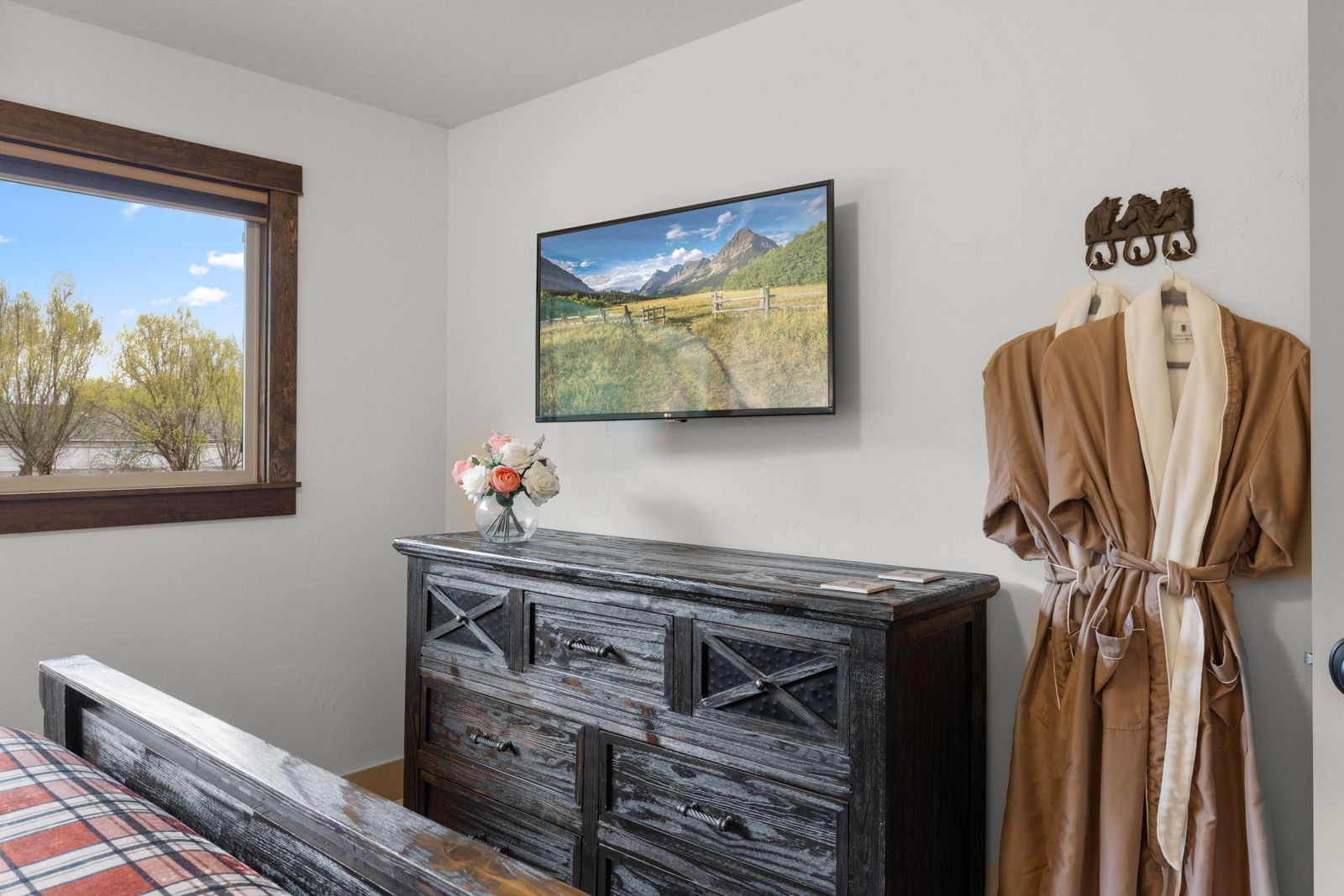 Bedroom corner with wall-mounted TV, black dresser, window, robe on a wall hook, and a neatly made bed edge