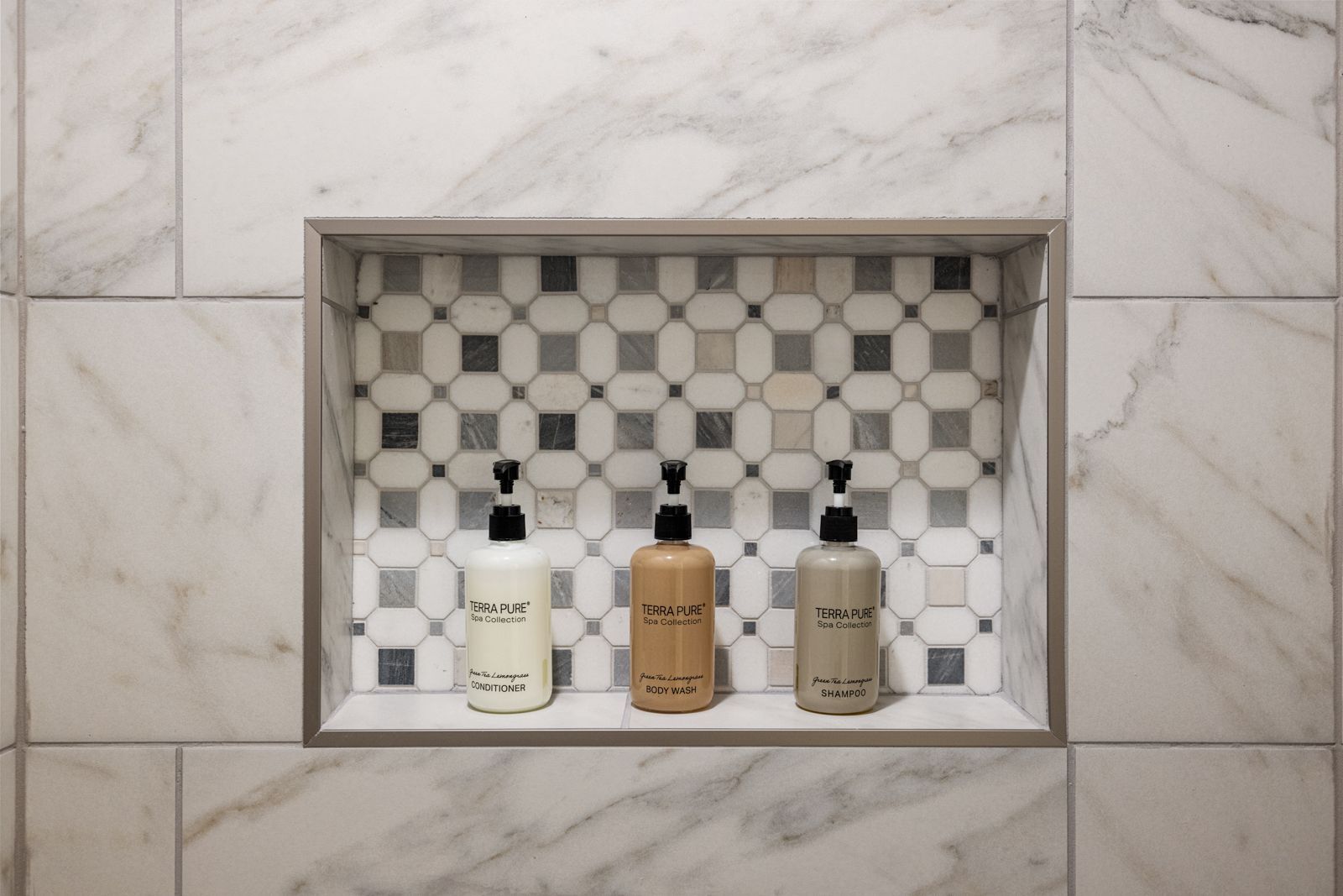Three soap dispensers in a tiled recessed bathroom niche with black-and-white mosaic backing.