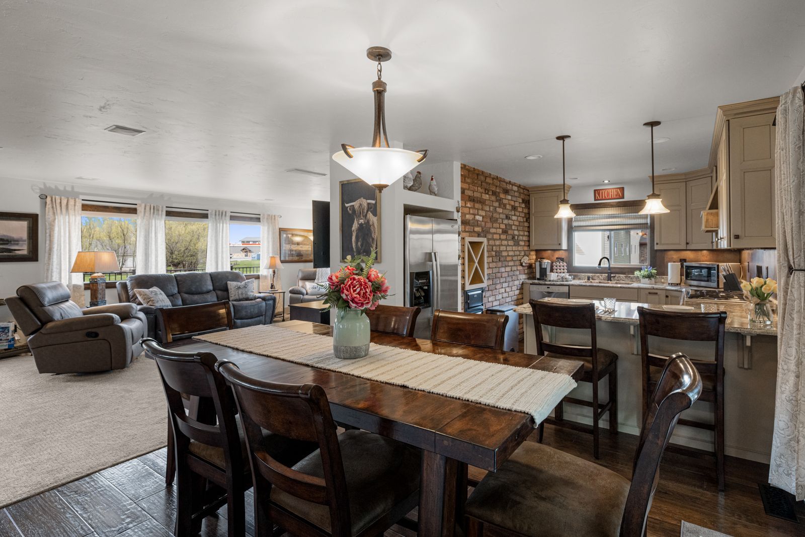 Open-plan kitchen and dining area with dark wood table, pendant lights, stone fireplace, and living room beyond