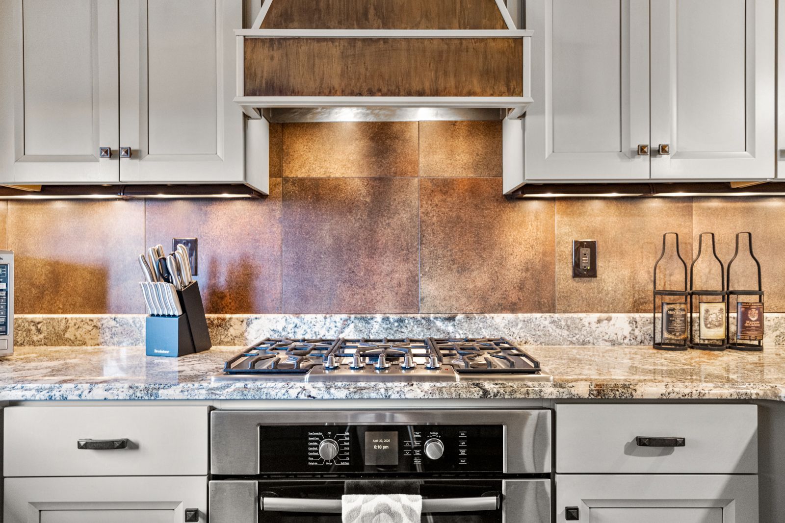 Modern kitchen stove with gray cabinets, granite countertop, and copper tile backsplash