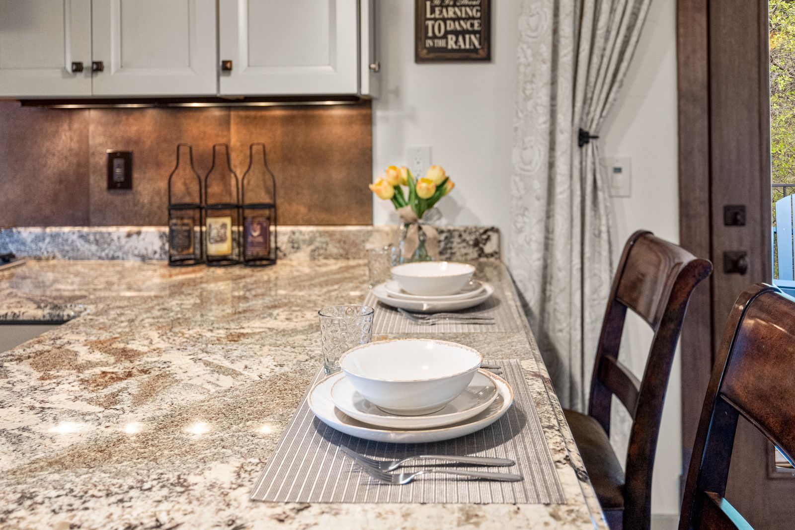Modern kitchen with granite island, set dining place settings, wine bottles, and flowers by the window