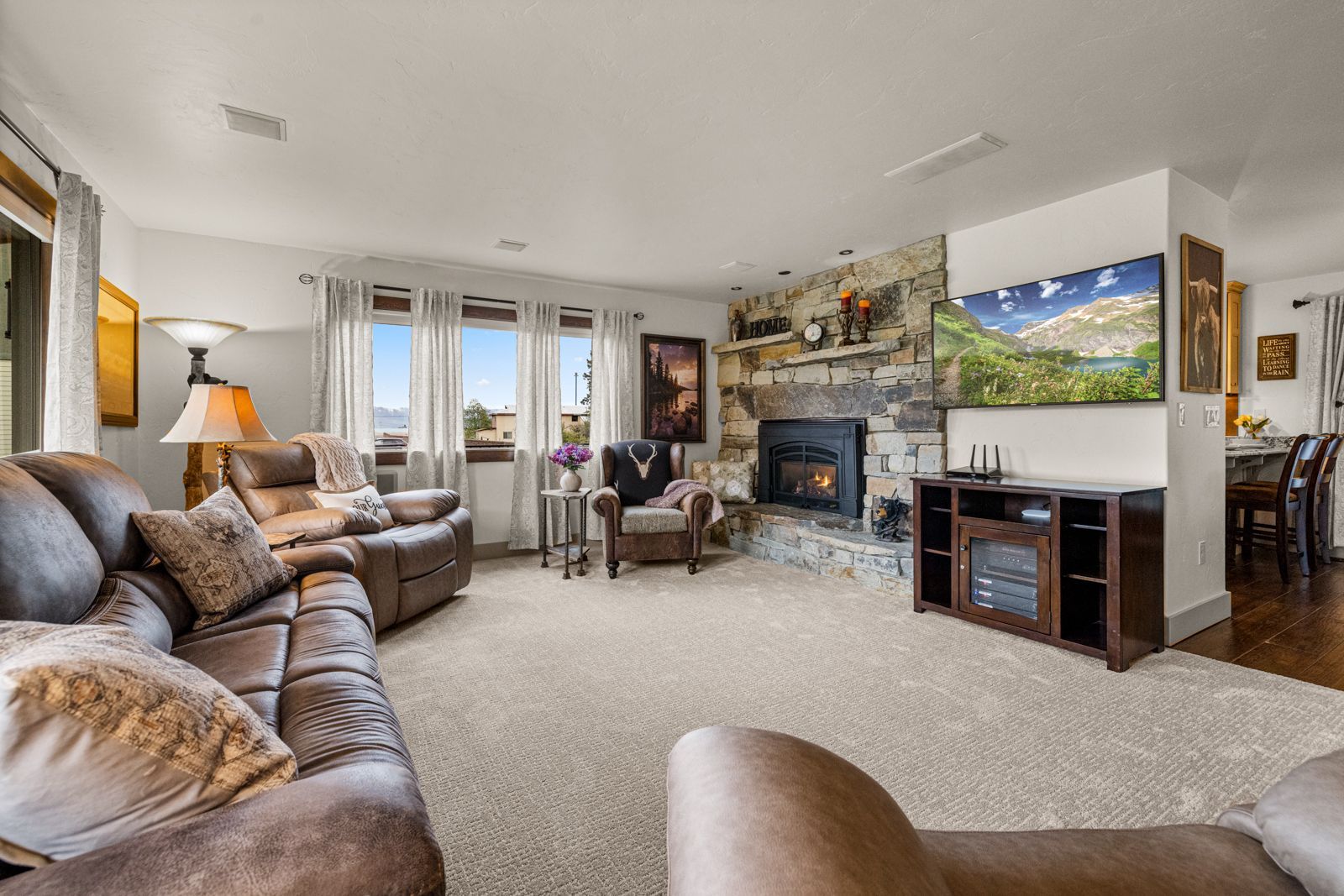 Bright living room with brown sofas, a fireplace, large windows, and a TV displaying a mountain landscape