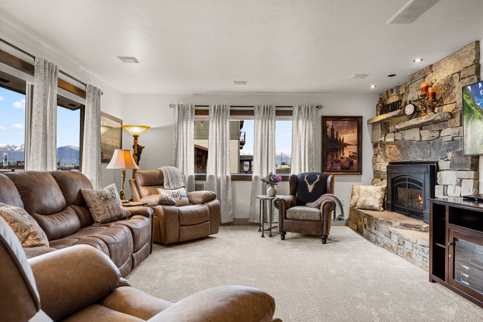 Bright living room with tan sofas, large windows, and a stone fireplace with TV.