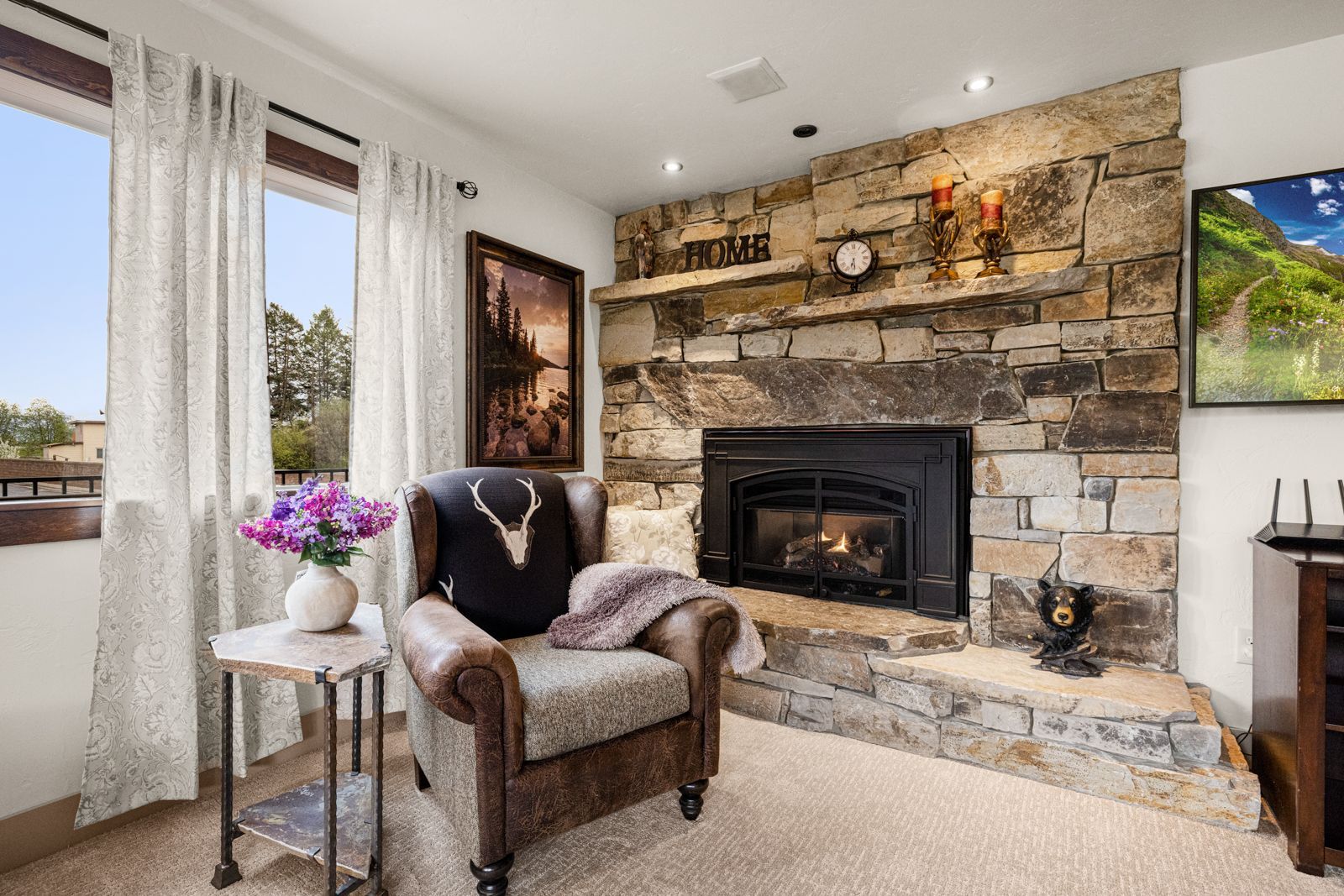 Cozy living room with stone fireplace, brown armchair, white curtains, and a vase of purple flowers.