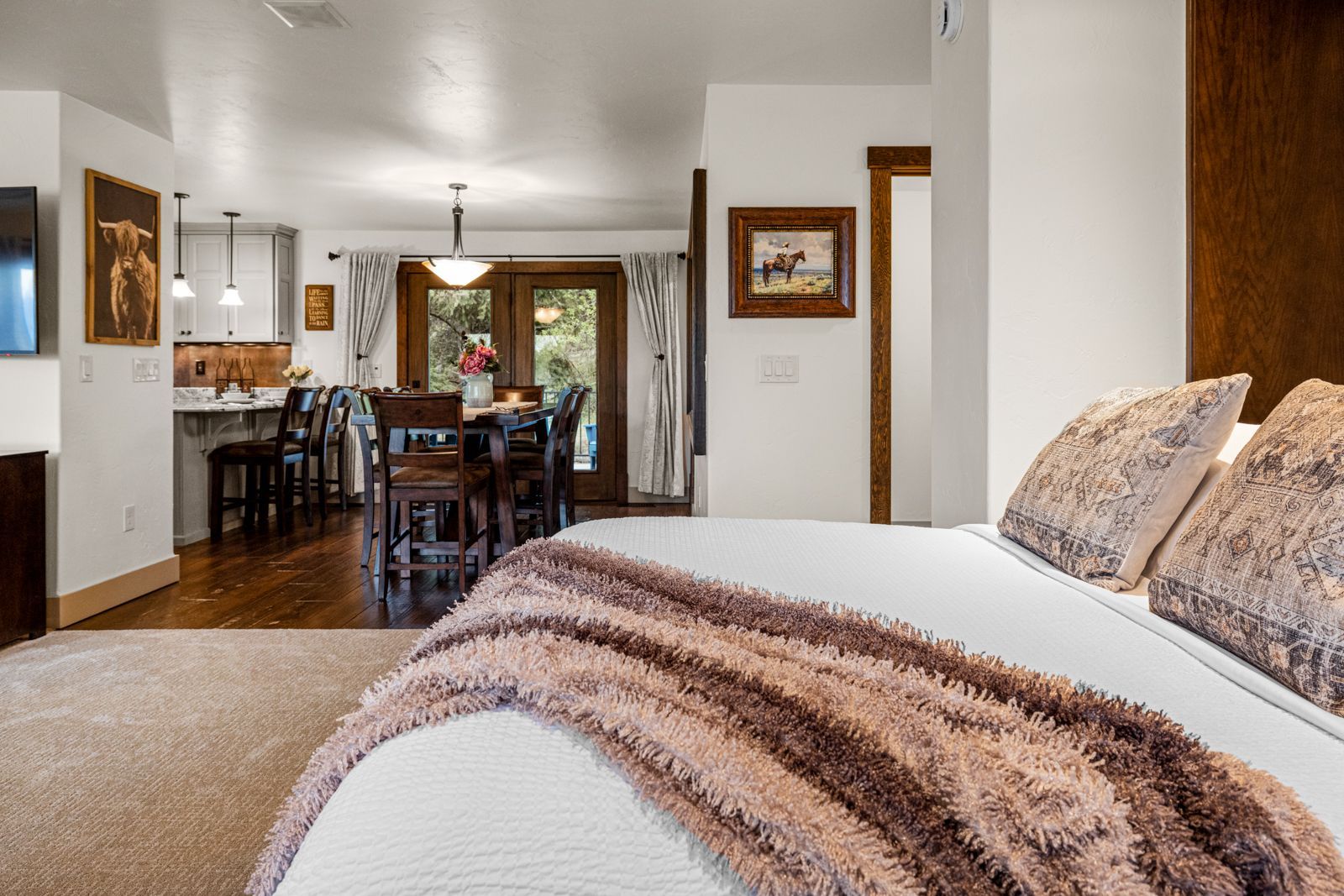 Bedroom with white bed and brown throw, looking into a dining room and kitchen through an open doorway