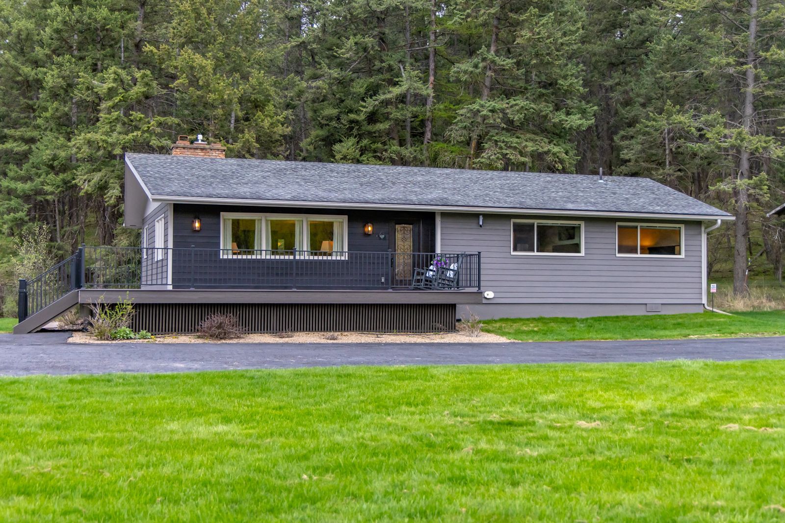 Gray cabin with a deck in a green lawn, set against a forest backdrop.