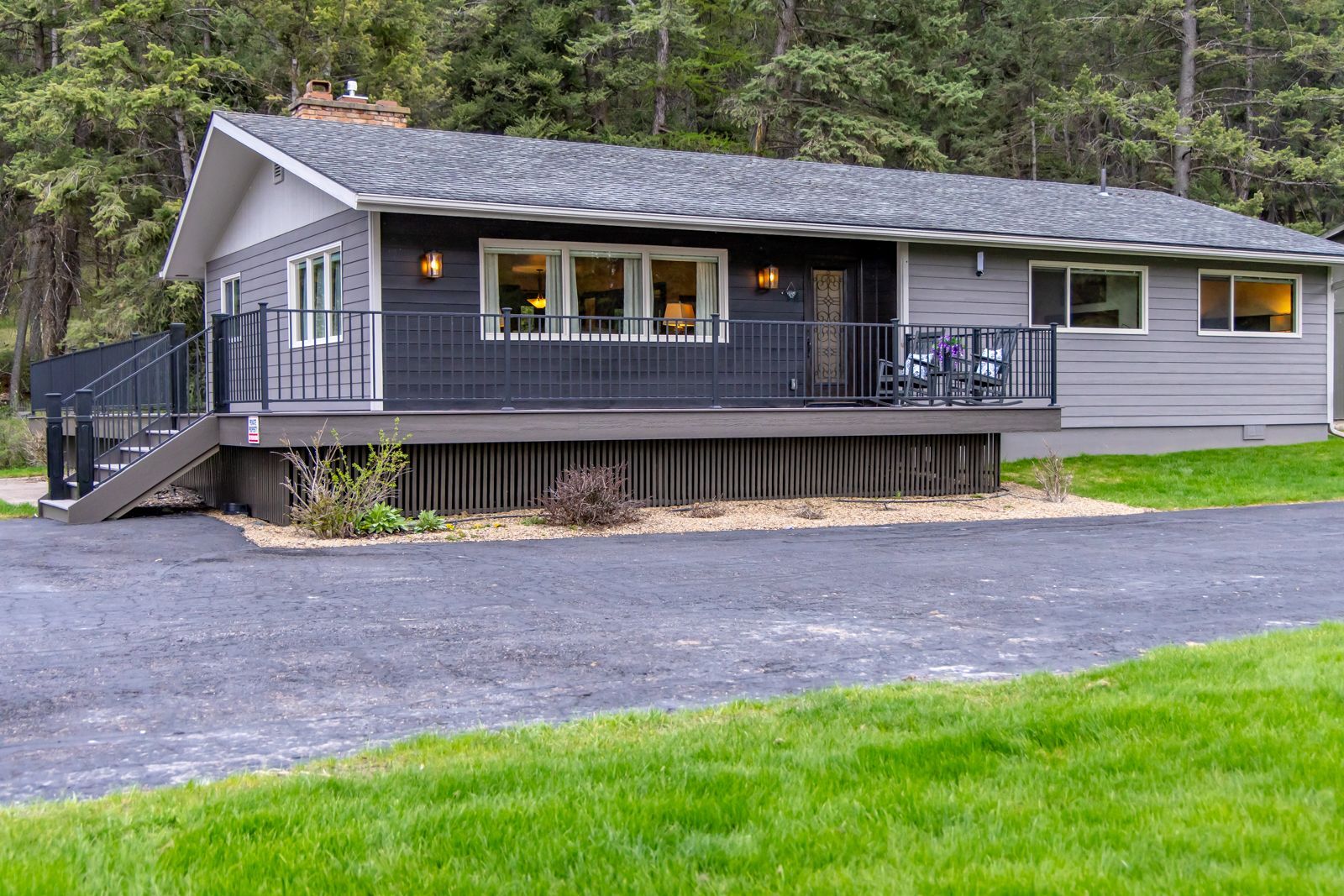 Gray cabin with a covered porch and deck, set beside a wooded area and gravel driveway