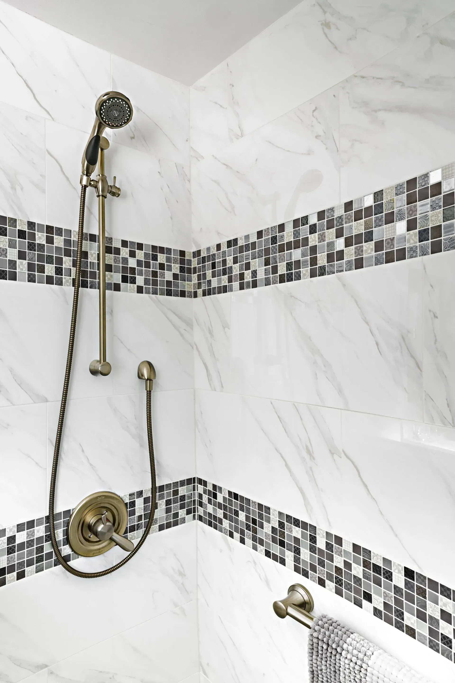 Modern shower with gold fixtures and gray mosaic tile accent in a marble-patterned bathroom in a modern kitchen with gray cabinets, island, pendant lights, exposed wood beams, and two barstools in Arlington Heights, IL.