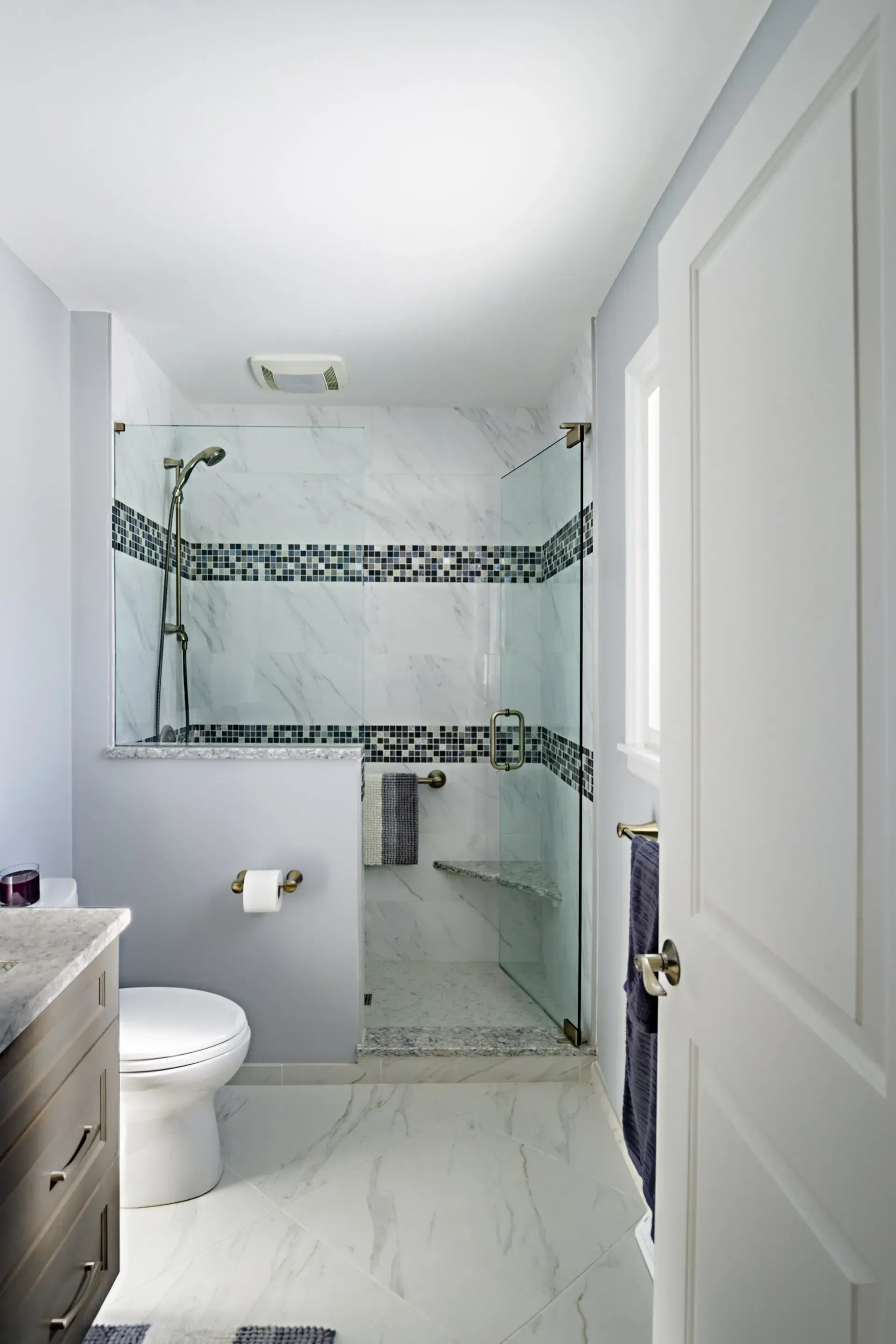 Bright bathroom with walk-in shower, white walls and floor, and decorative tile bands. in a modern kitchen with gray cabinets, an island, pendant lights, exposed wood beams, and two barstools in Arlington Heights, IL.