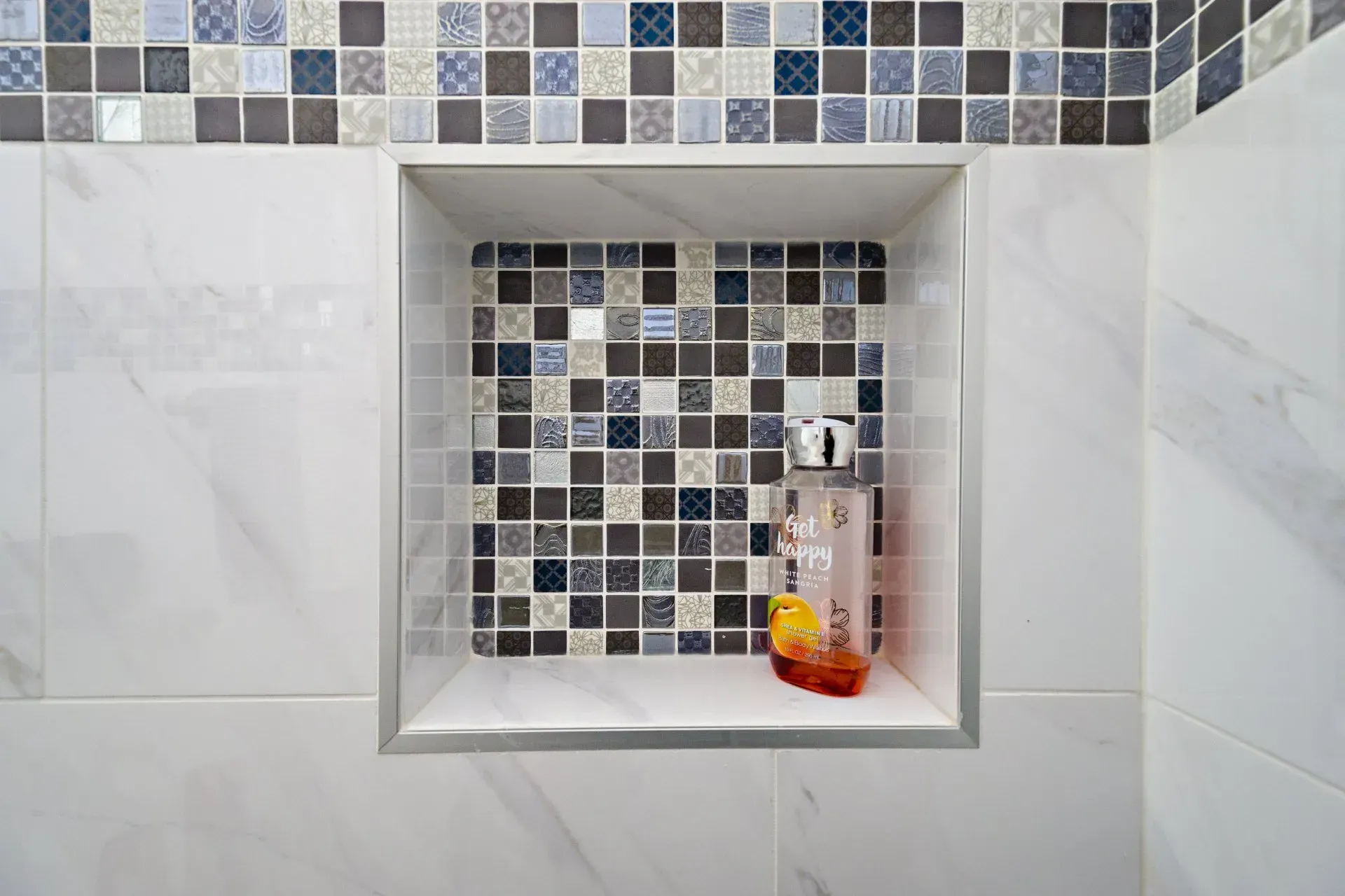 Bathroom shower niche with gray and blue mosaic tile and a soap dispenser with orange fruit decor in a modern kitchen with gray cabinets, island, pendant lights, exposed wood beams, and two barstools in Arlington Heights, IL.