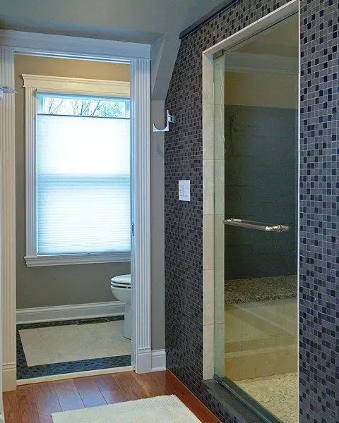 Bathroom doorway beside a mosaic-tiled shower, with a windowed toilet alcove in the background. In a modern kitchen with gray cabinets, an island, pendant lights, exposed wood beams, and two barstools in Arlington Heights, IL.
