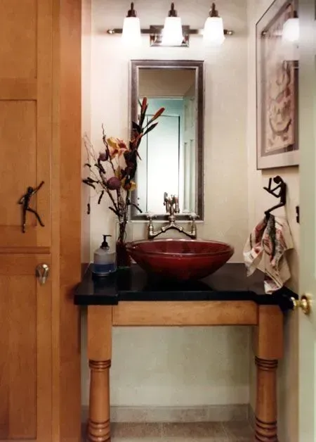 Small bathroom vanity with red bowl sink, mirror, wall lights, and floral decor in a modern kitchen with gray cabinets, island, pendant lights, exposed wood beams, and two barstools in Arlington Heights, IL.