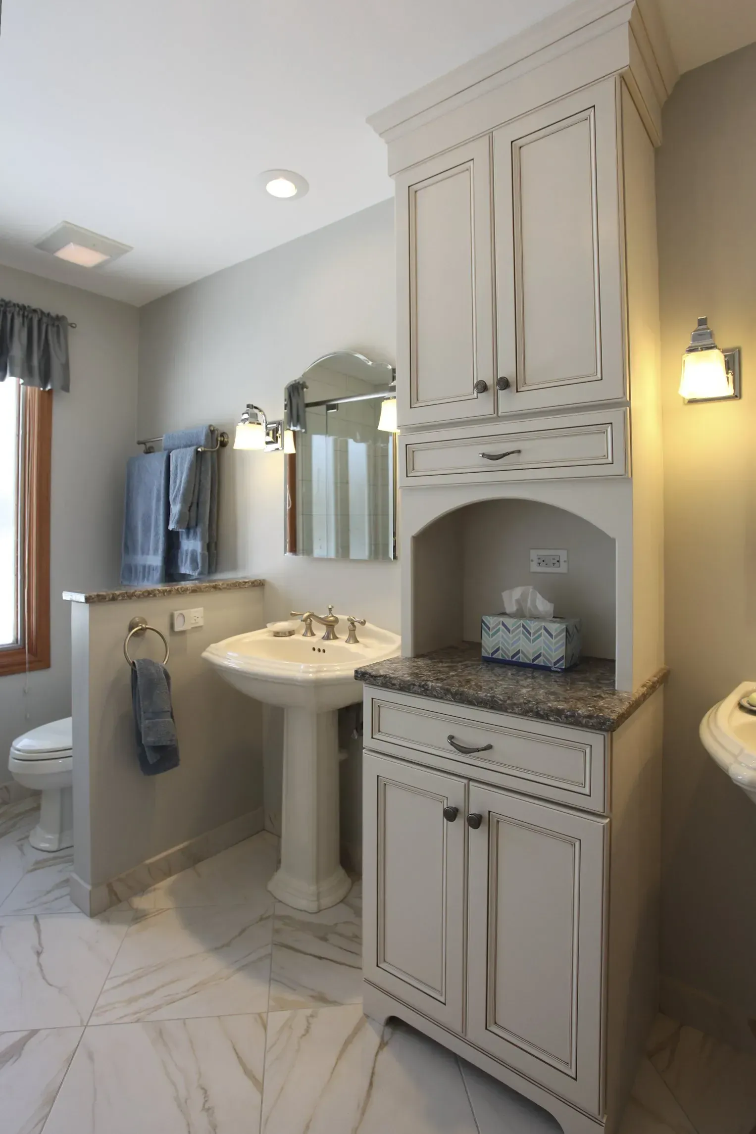Bright bathroom with white vanity, pedestal sink, mirror, and built-in cabinet under warm wall light in Modern kitchen with gray cabinets, island, pendant lights, exposed wood beams, and two barstools in Arlington Heights, IL.