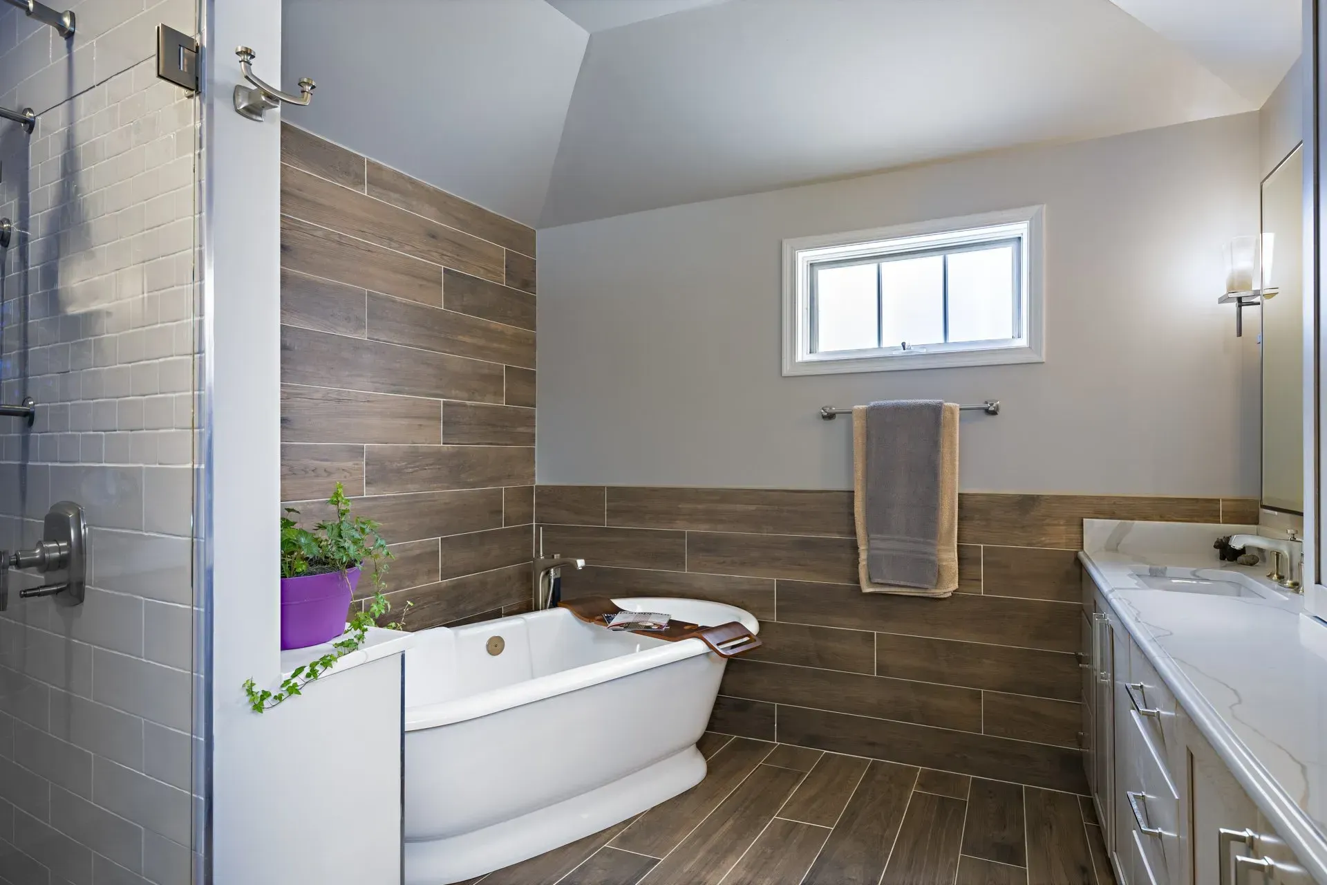 Modern bathroom with freestanding tub, tiled walls, vanity, and small window with towel rack in Modern kitchen with gray cabinets, island, pendant lights, exposed wood beams, and two barstools in Arlington Heights, IL.