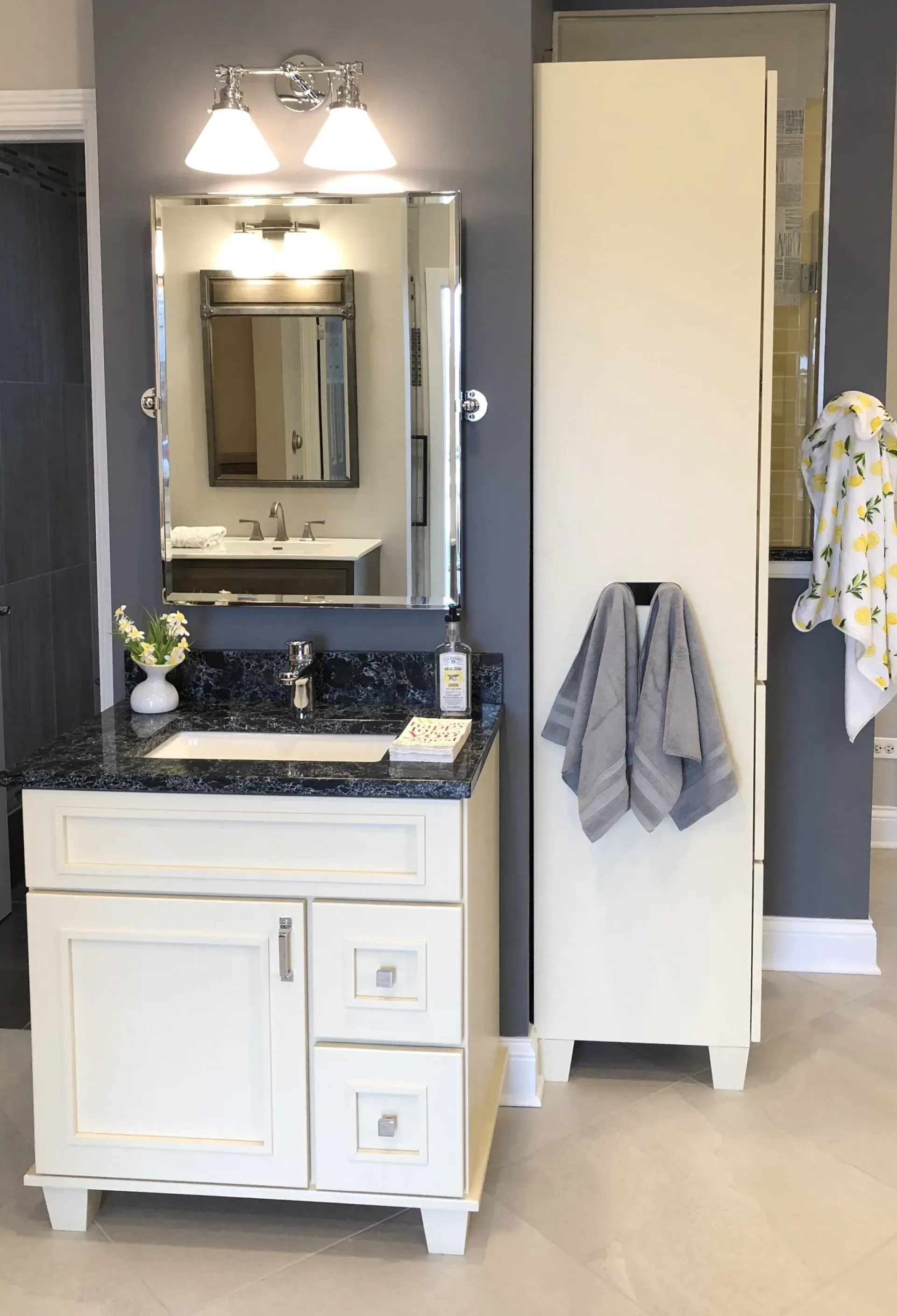 Bathroom vanity with dark blue countertop, white cabinets, mirror, sink, and a towel rack with hanging towels in a modern kitchen with gray cabinets, island, pendant lights, exposed wood beams, and two barstools in Arlington Heights, IL.