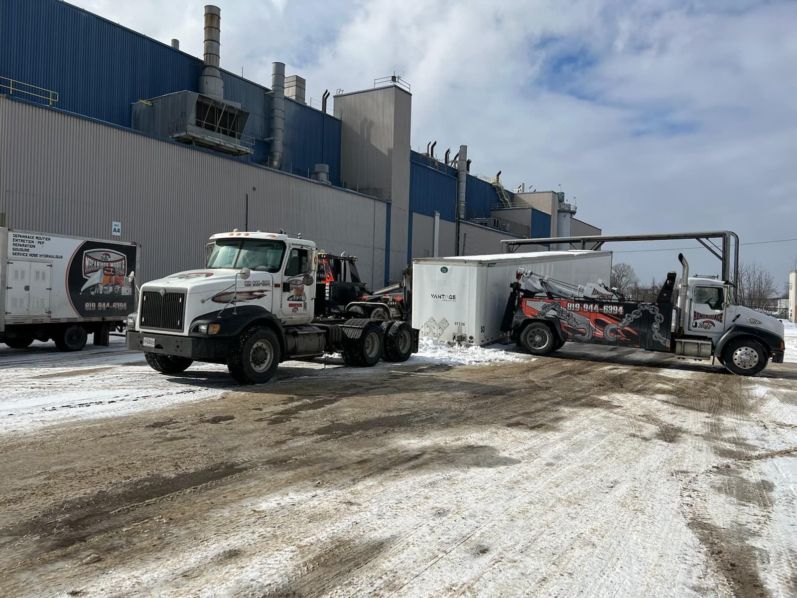 Trois camions de service blancs garés à l'extérieur d'un grand bâtiment industriel par une journée enneigée.