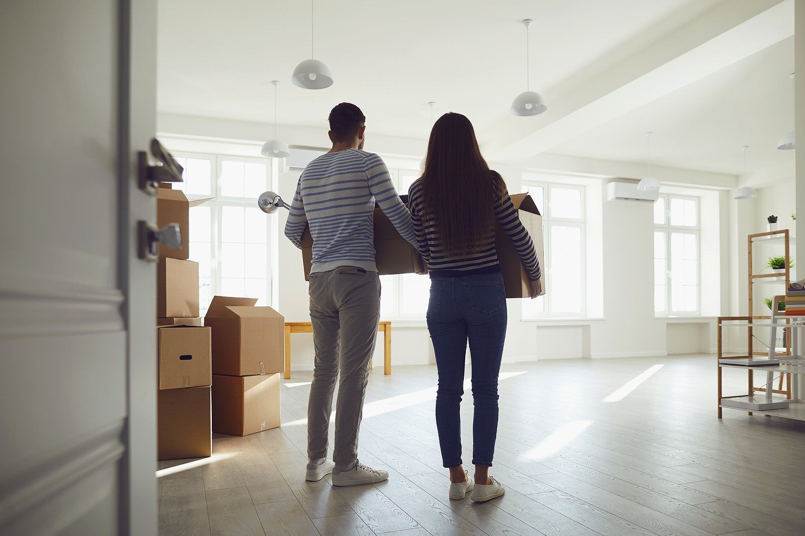 Two people stand in a bright, empty room with moving boxes and sunlight coming through large windows.