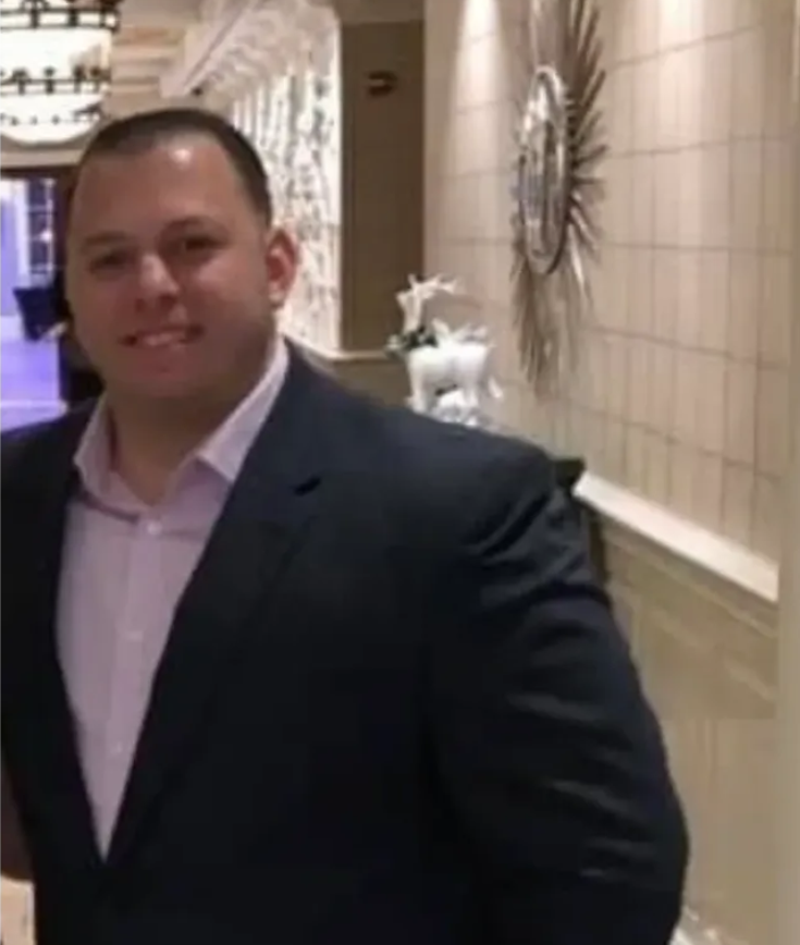 A man in a suit and a pink shirt is standing in a hallway.