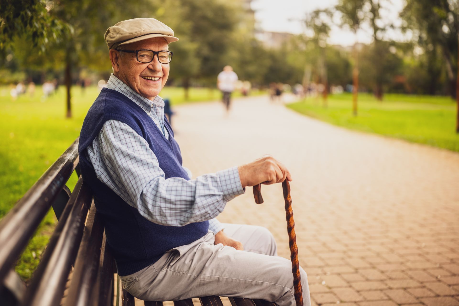 Elderly man sitting on a park bench, smiling, holding a cane. He wears a hat, vest, and light-colored pants.