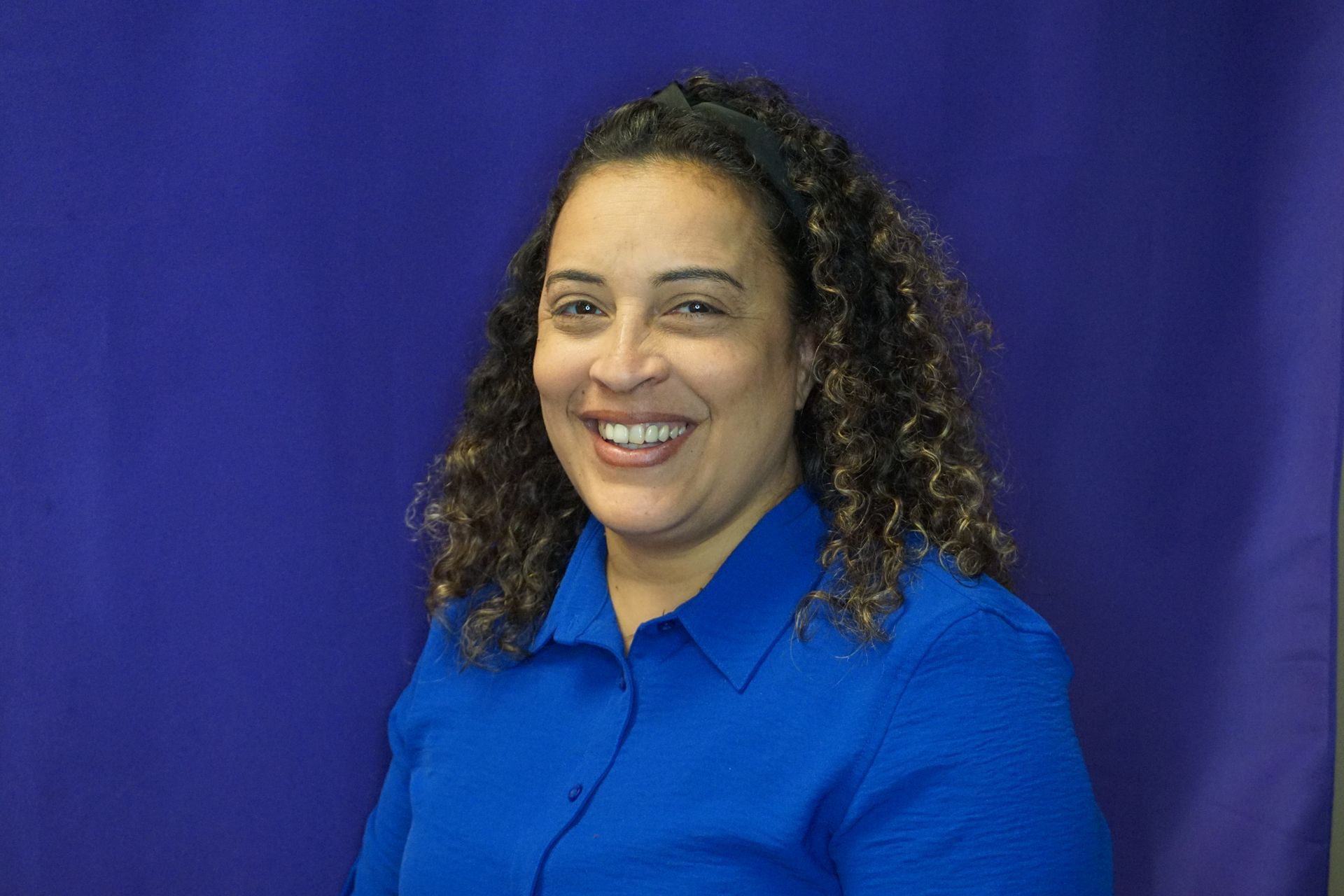 A woman in a blue shirt is smiling in front of a purple background.