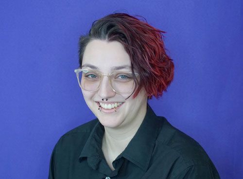 A woman with red hair and glasses is smiling in front of a blue background.