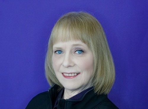 A woman with blonde hair and blue eyes is smiling in front of a purple background.